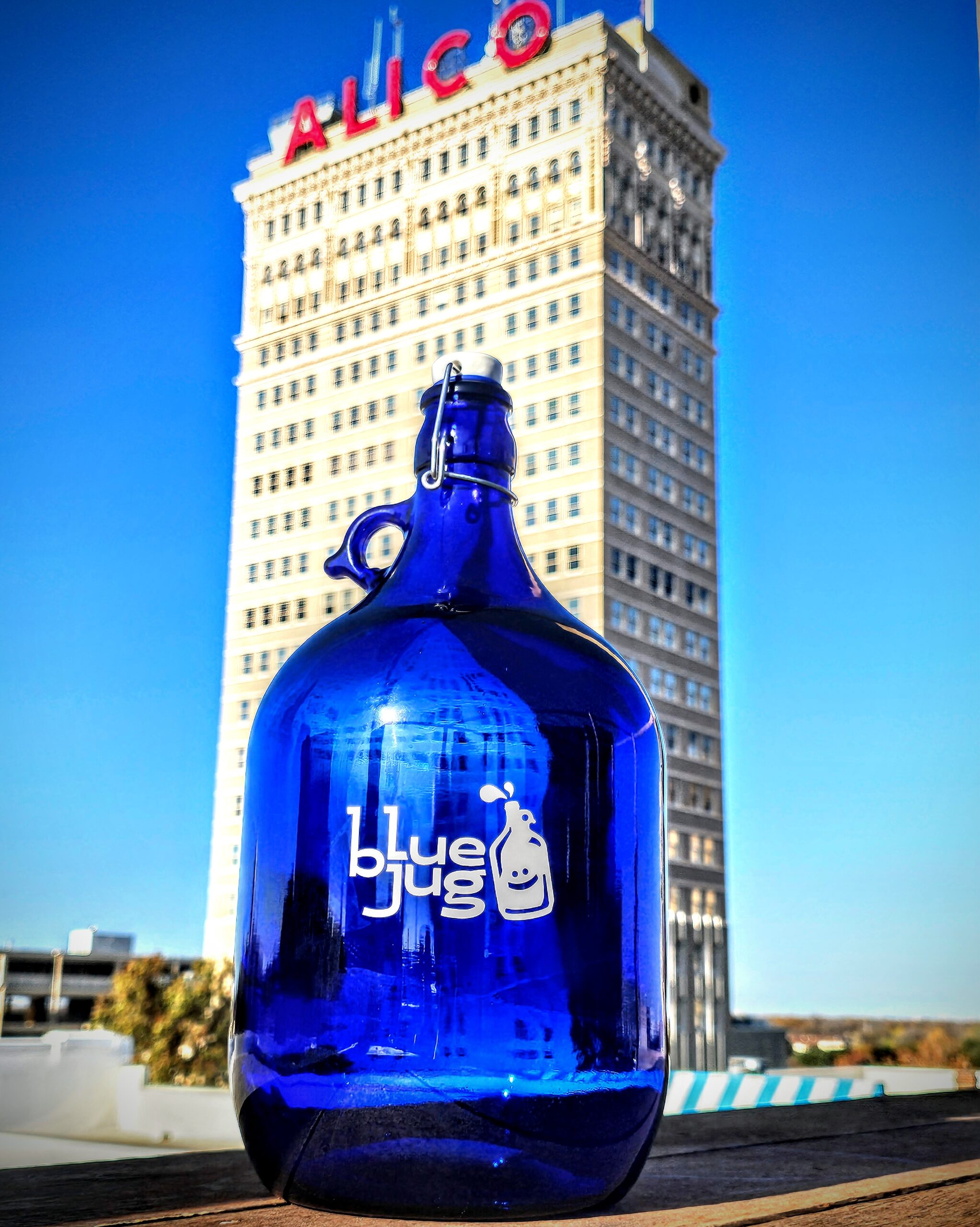 A dark blue bottle placed near the ALICO Building with the "Blue Jug Waco" logo on it